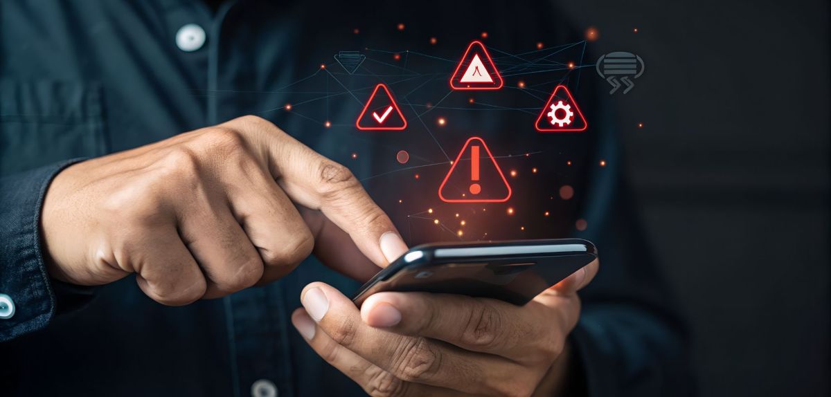 Person hält ein Smartphone in der Hand. Darüber wird ein Warn-Symbol angezeigt.