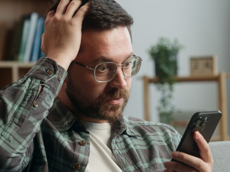 Mann hält ein Handy in der Hand und fasst sich verwundert an den Kopf.