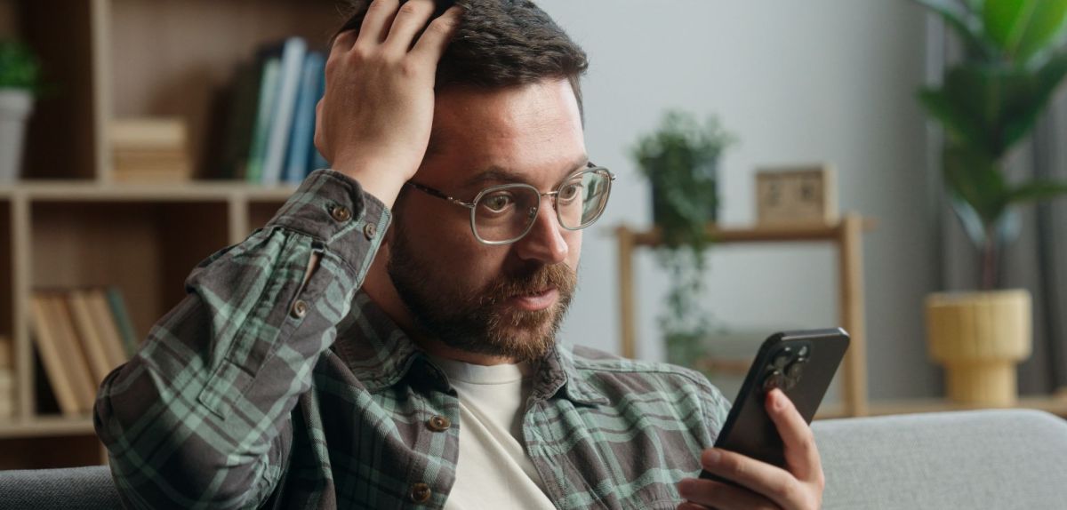 Mann hält ein Handy in der Hand und fasst sich verwundert an den Kopf.