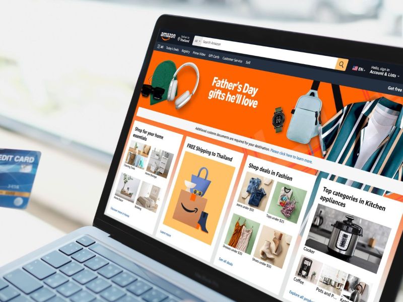 Auf einem Laptop wird die Amazon-Seite angezeigt. Im Vordergrund hält eine Person eine Bankkarte in der Hand.