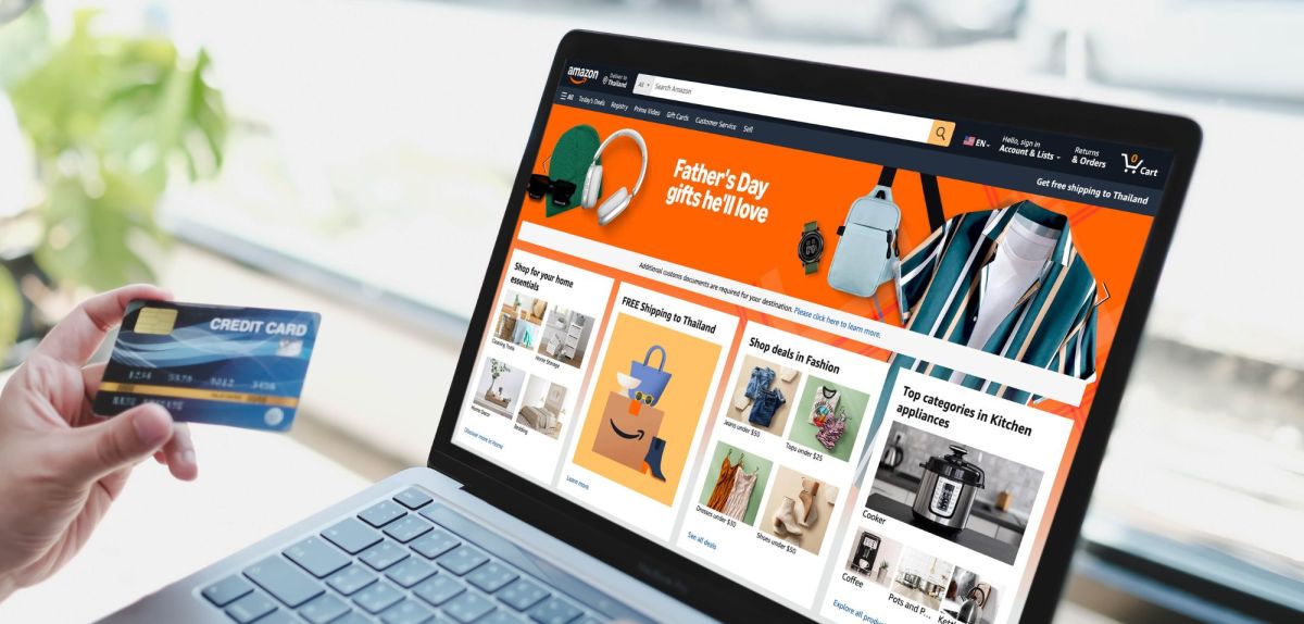 Auf einem Laptop wird die Amazon-Seite angezeigt. Im Vordergrund hält eine Person eine Bankkarte in der Hand.