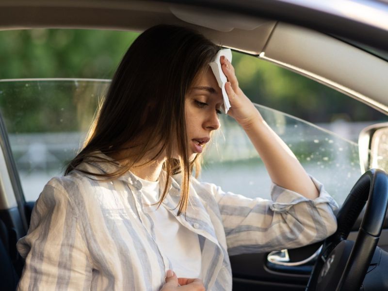 Frau sitzt bei Hitze im Auto und schwitzt stark. Sie hält sich ein Tuch an Kopf.
