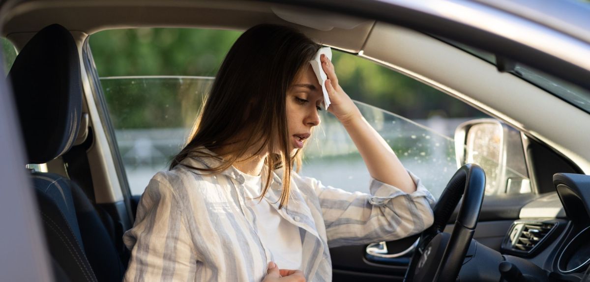 Frau sitzt bei Hitze im Auto und schwitzt stark. Sie hält sich ein Tuch an Kopf.