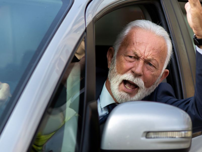 Mann sieht aus dem Autofenster mit wütendem Gesicht und hebt erbost den Arm.