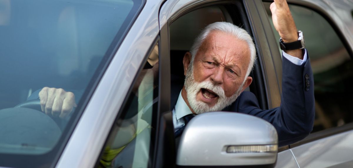 Mann sieht aus dem Autofenster mit wütendem Gesicht und hebt erbost den Arm.