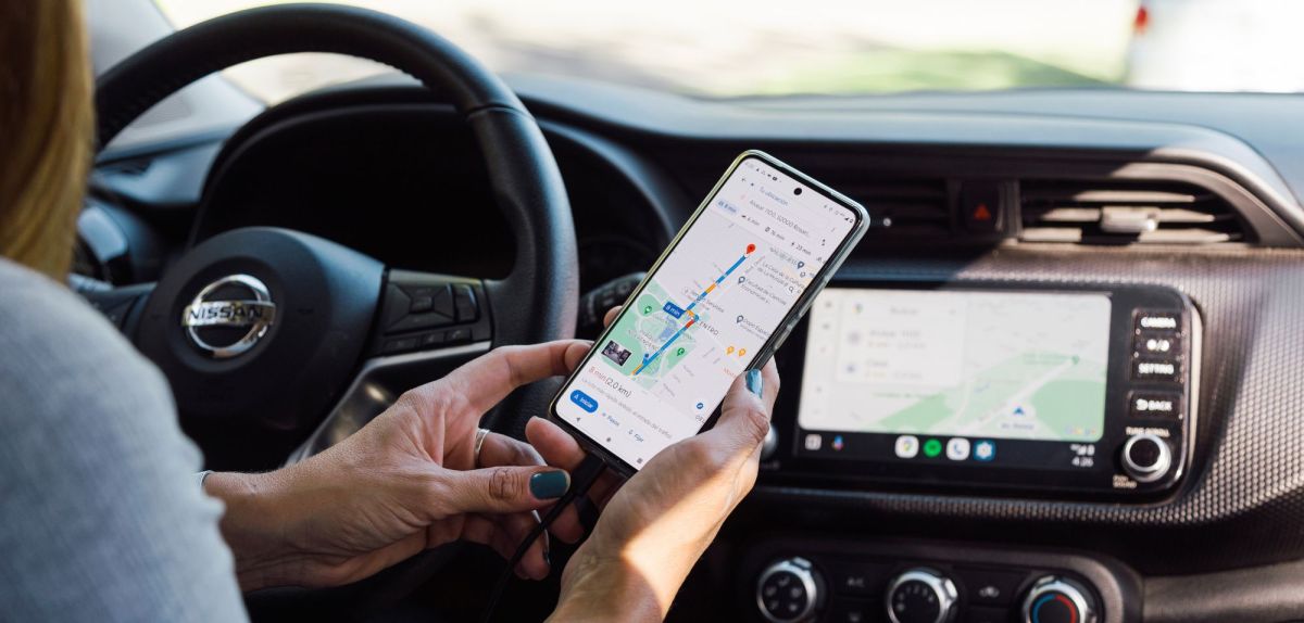 Person sitzt auf dem Fahrersitz eines Autos. Die Person hält ein Handy in der Hand. Auf dem Display wird Google Maps angezeigt.