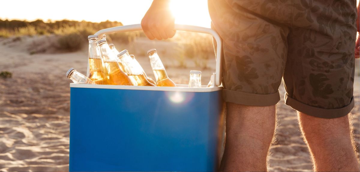 Person steht mit einer Kühlbox in der Hand an einem Strand.