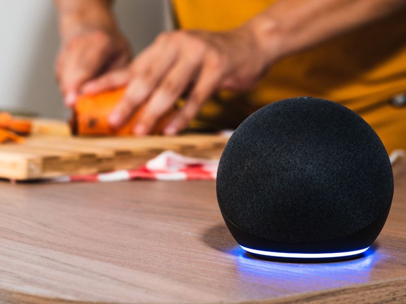 Amazon Echo Dot steht auf einen Tisch. Im Hintergrund ist eine Person zu sehen, die Brot schneidet.