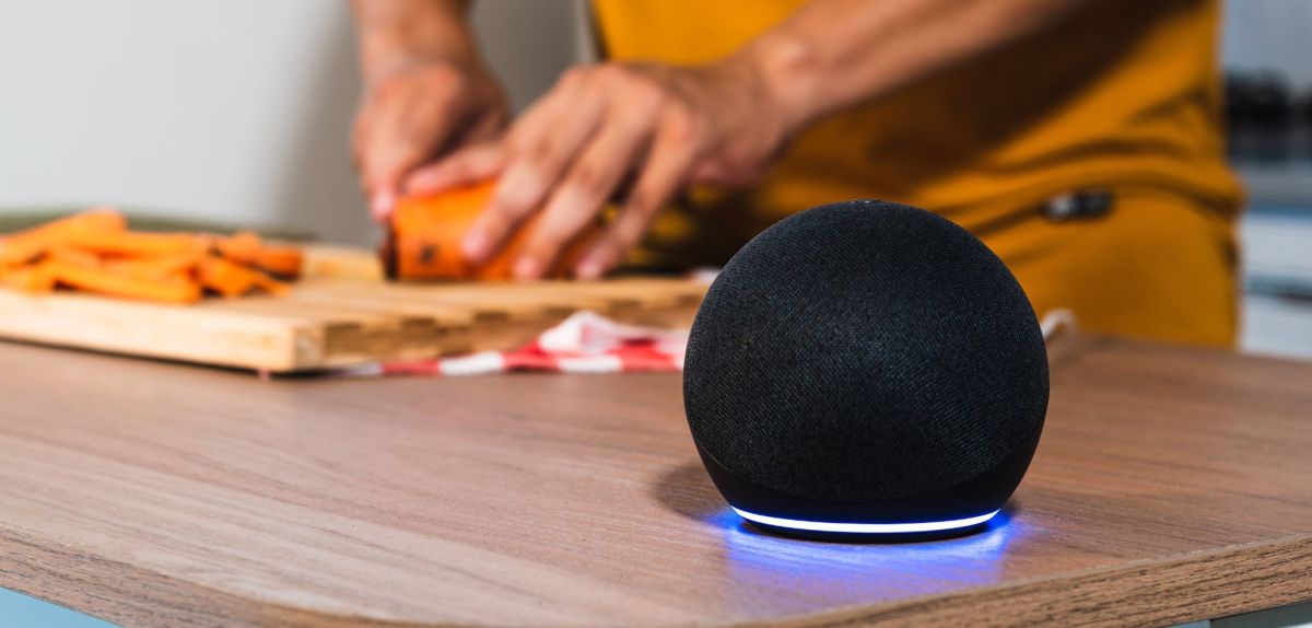 Amazon Echo Dot steht auf einen Tisch. Im Hintergrund ist eine Person zu sehen, die Brot schneidet.
